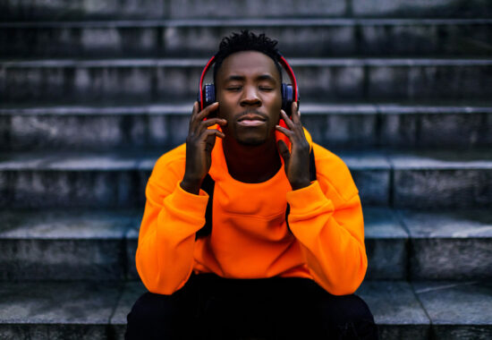 african-american man in wireless headphones listening music african-american man in stylish orange hoodie sweatshirt in wireless headphones listening music and enjoying music on background of stairs